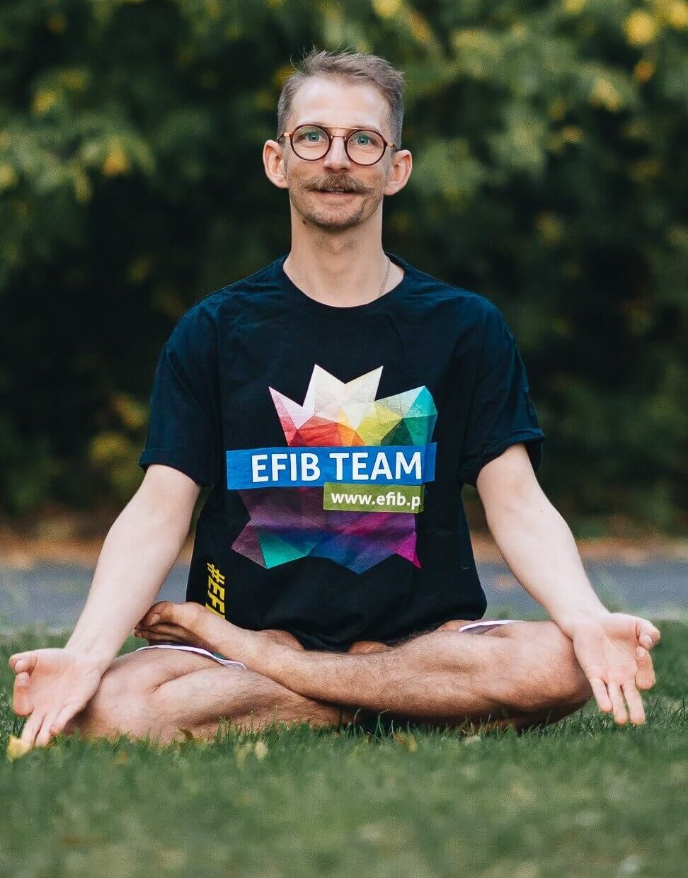 Trener w koszulce z logo efib, nauczyciel jogi w asanie siedzi na łące.