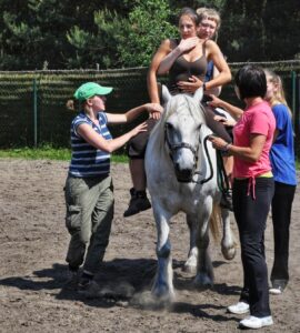 grupa osób wokół konia asystuje podczas hipoterapii. Na koniu pacjent i podtrzymujący go hipoterapeuta. kurs hipoterapii