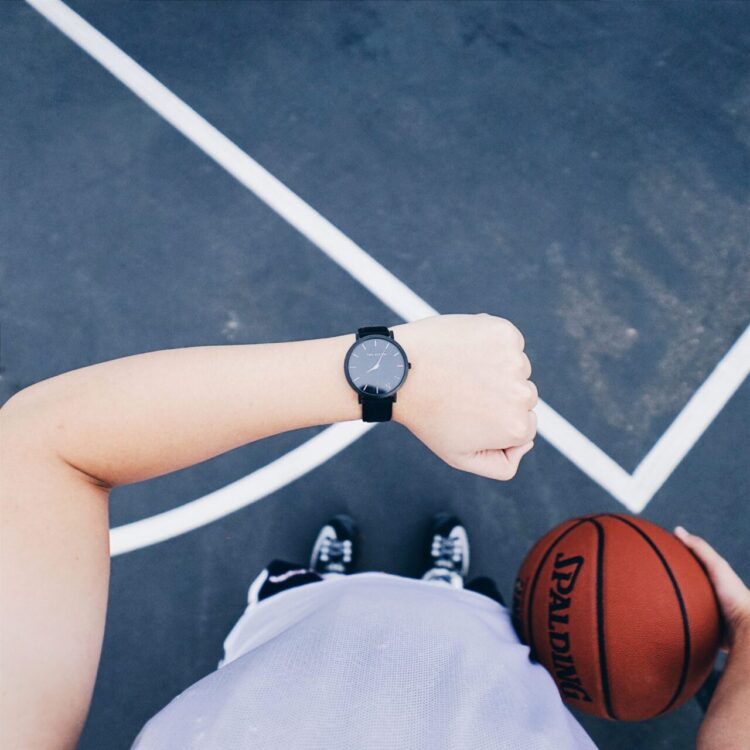 Sylwetka sportowca, który patrzy na godzinę na zegarku. Aktywność fizyczna ruch. W drugiej dłoni piłka. W tle boisko do koszykówki.